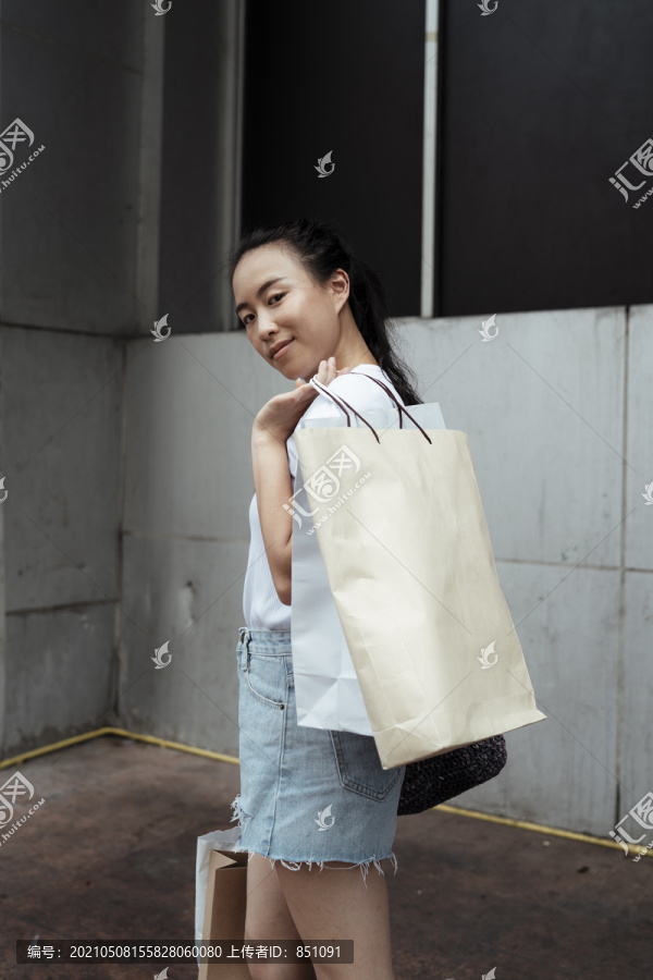 年轻的泰国亚裔女子，穿着白色衬衫和牛仔裤短裤，背着许多购物袋。