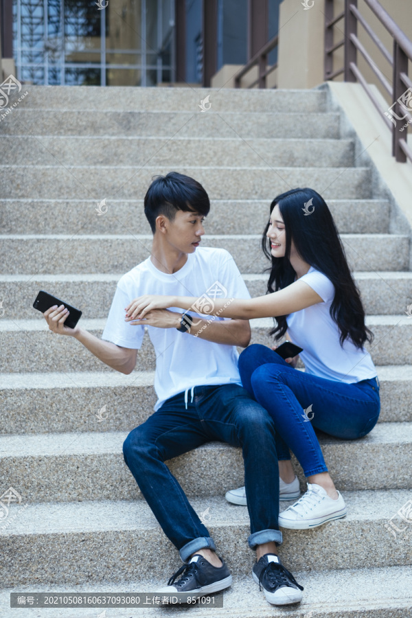 一对穿着白色t恤的情侣坐在鹅卵石楼梯上，互相取笑，展示智能手机屏幕。