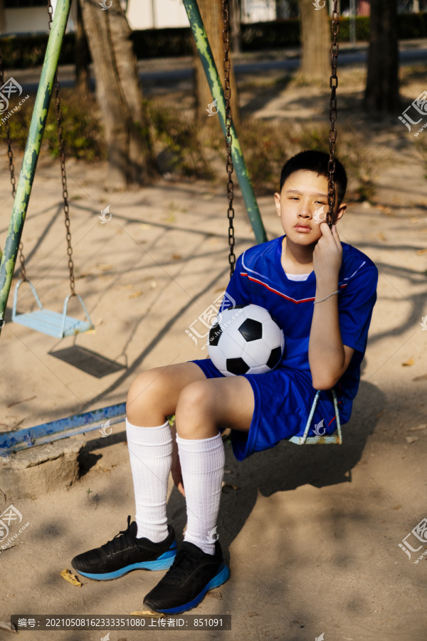 一个泰国小足球运动员坐在操场的秋千上拿着球。