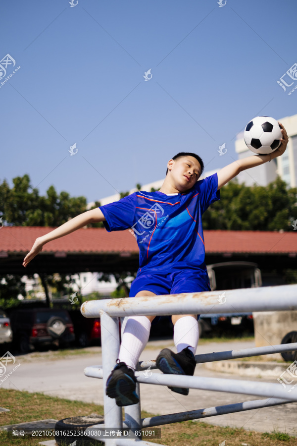 一个疯狂的男孩以危险的姿势坐在扶手上踢足球。以蓝天为背景。