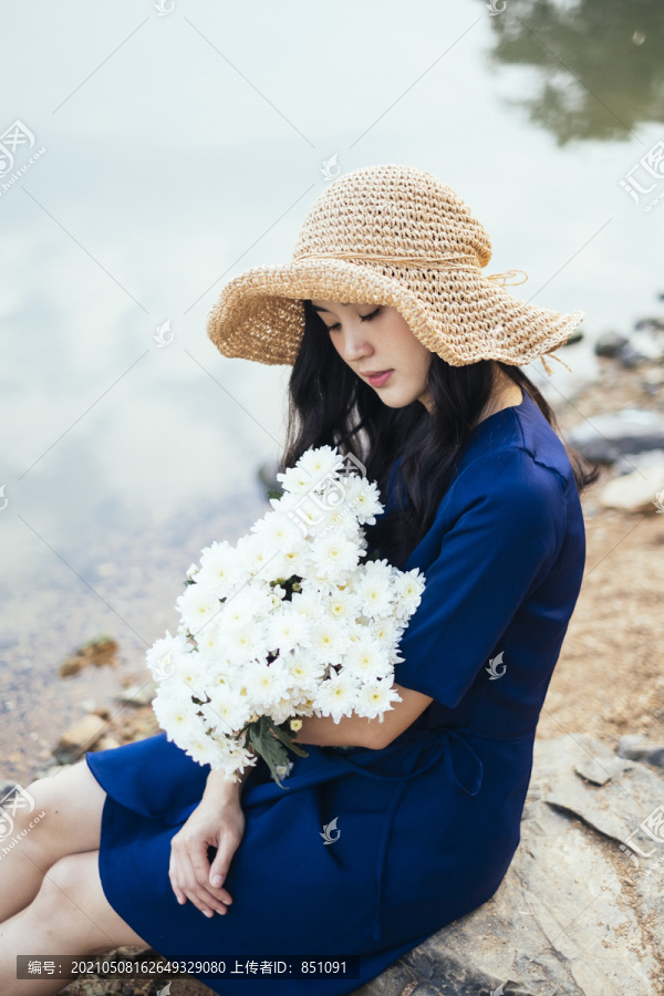 一个穿着深蓝色裙子的可爱女孩坐在湖边，爱上了怀中的白花。