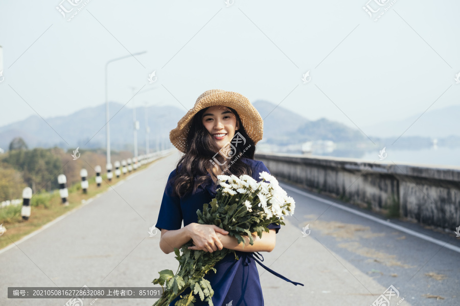 身着深蓝色连衣裙的可爱女孩微笑着拥抱大坝路上的白花的中写真。