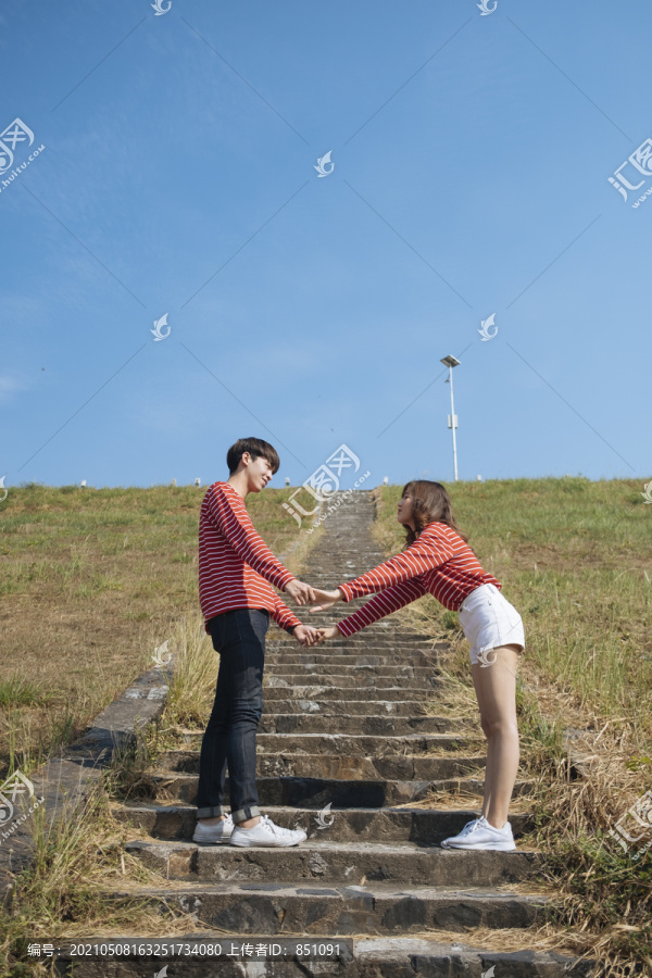 一对年轻的情侣站在石阶上，在蓝天下互相牵手。