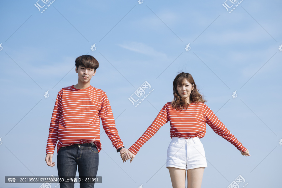 以蓝天为背景，一对年轻的情侣站在一起，手牵着手。