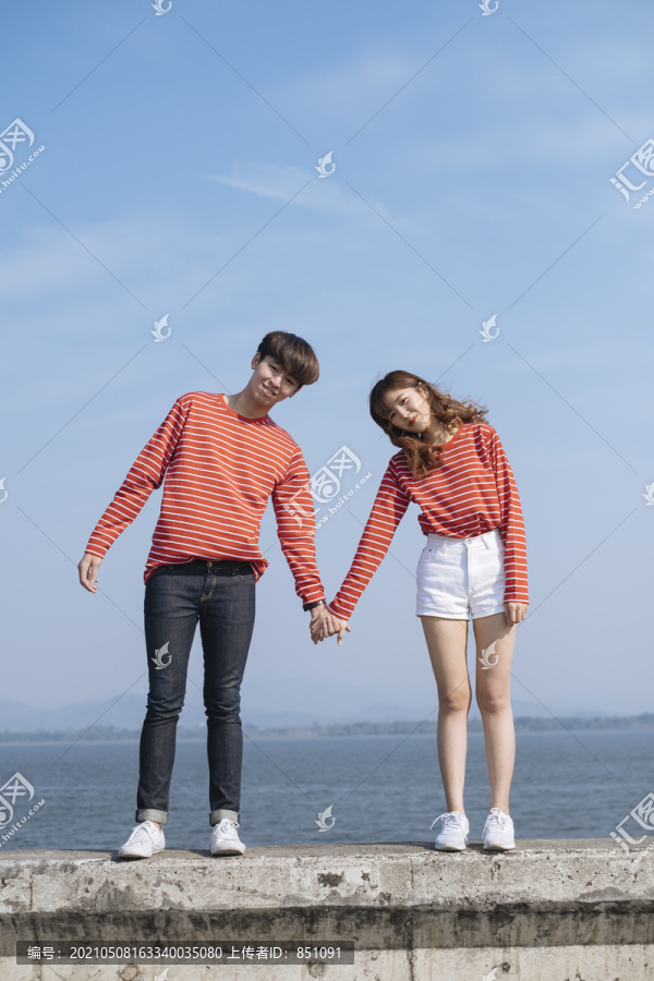 一对年轻的夫妇站在大坝的路障上，手牵着手，仰着头看海景。