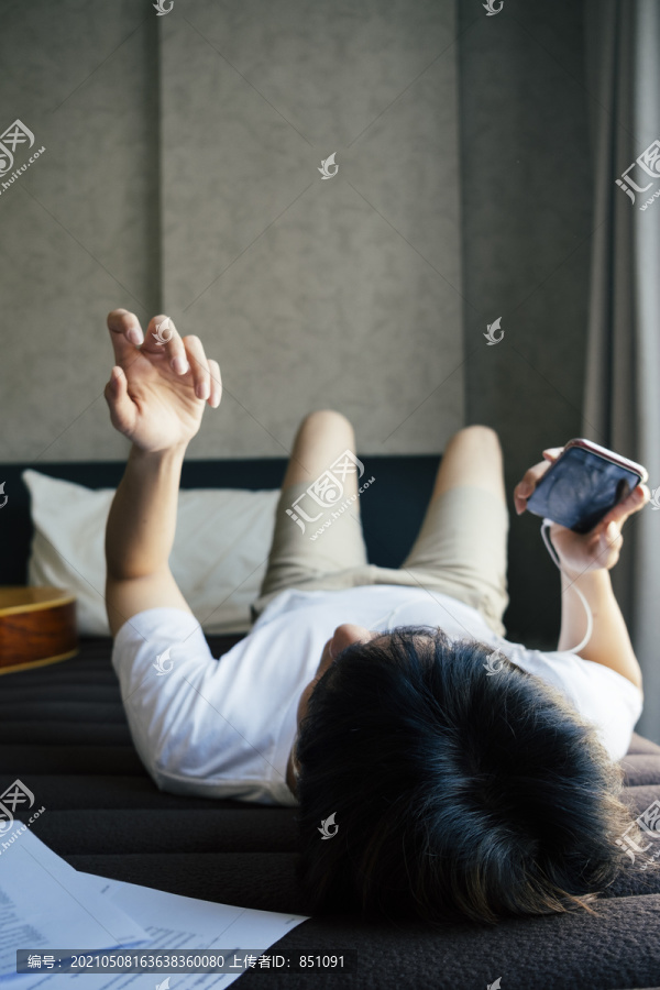年轻的泰国吉他手面朝上躺在床上听智能手机里的歌曲，练习空气吉他。