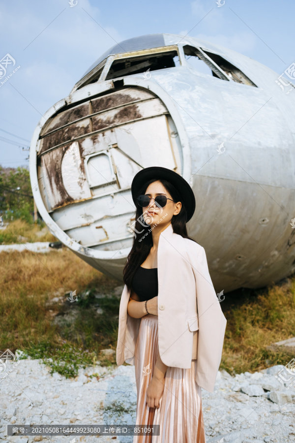 黑色长发女孩穿着粉色西装，戴着黑色帽子和墨镜站在废弃地区的废墟飞机前。