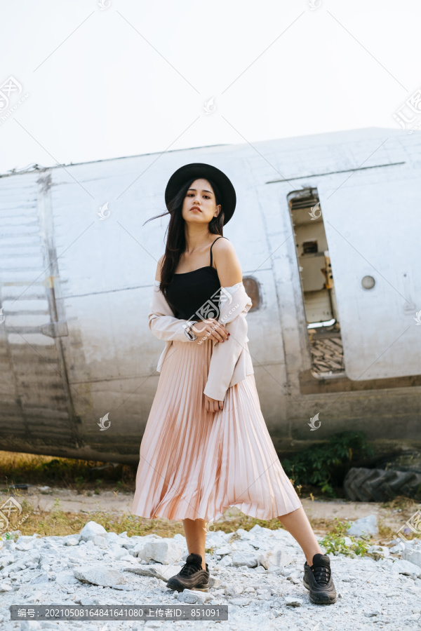 一位身穿黑色和粉色衣服的长发女子站在失事飞机前，穿着粉色西装露出肩膀，闭上嘴。