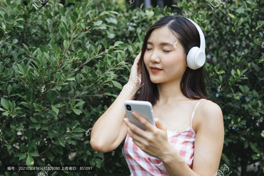 戴着耳机的美丽年轻女子在绿树旁的花园里闭着眼睛微笑着听音乐。