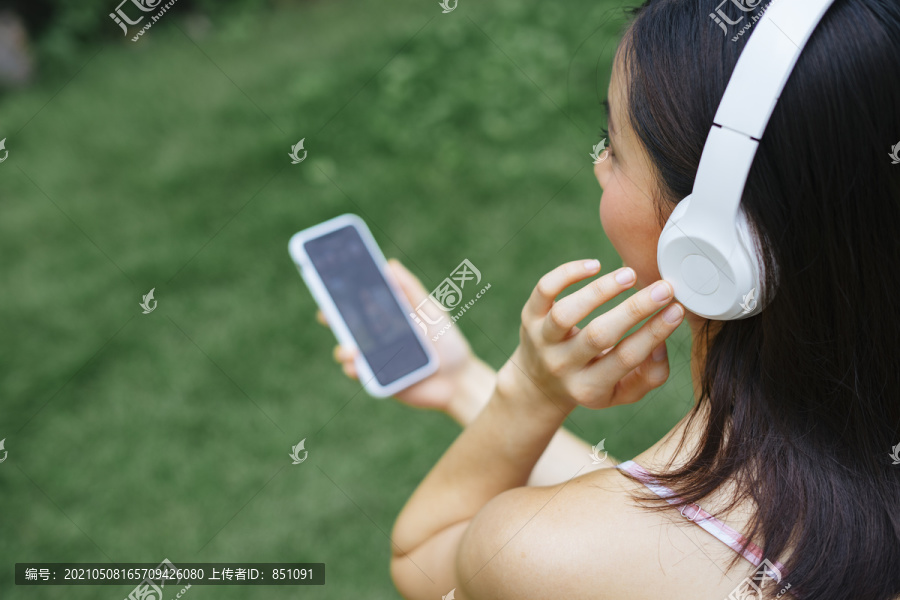 肩顶视图-阿西娜女孩用白色无线耳机听音乐，手持智能手机控制音乐平台。