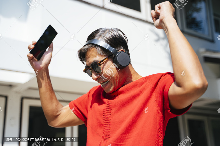 在阳光明媚的日子里，穿着红色t恤的老人在家里欣赏移动应用程序的音乐。