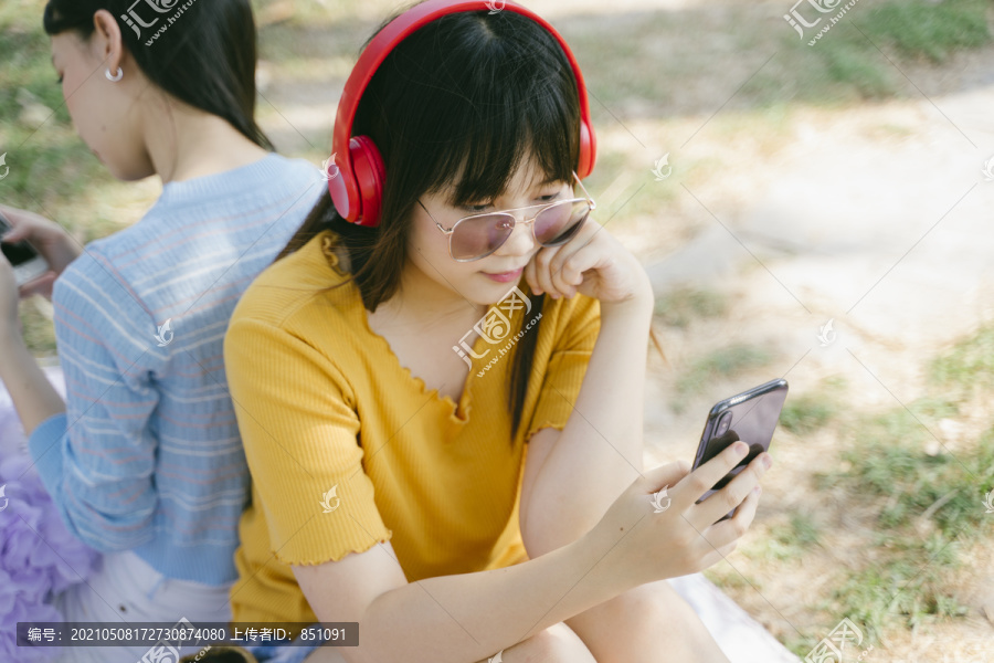 两个女孩背靠背地坐在地板上，听着智能手机应用程序里的歌曲，在社交媒体上旅行。