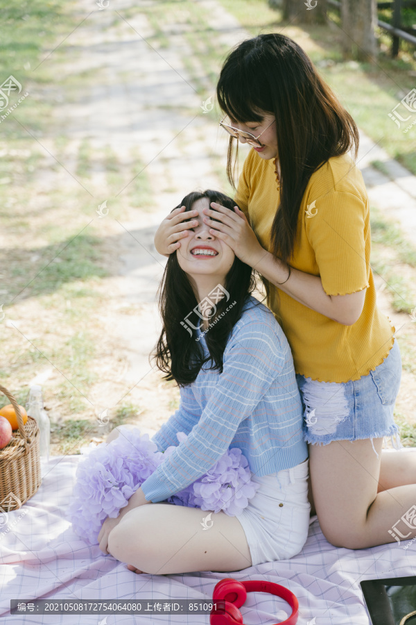 野餐时两个女孩在公园里玩耍，黄色t恤女孩闭上蓝色t恤女孩的眼睛。
