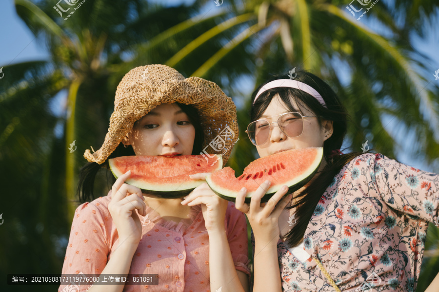 傍晚日落时，两个女孩站在沙滩上吃西瓜。