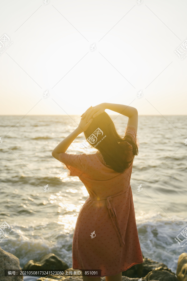 长发女孩站在海边的岩石上，悠闲地看着夕阳。