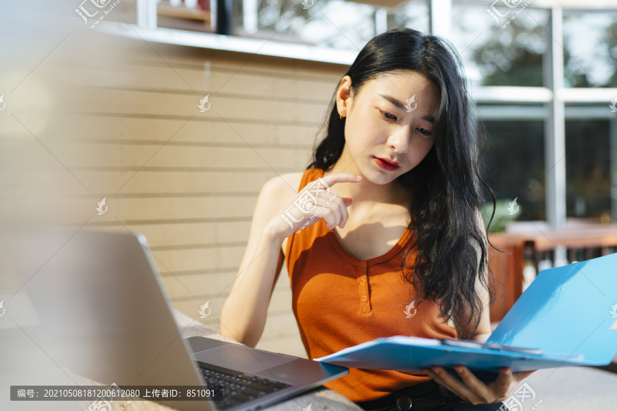 性感的亚洲女人穿着橙色衬衫在家里工作拿着蓝色的剪贴板文件看着笔记本电脑。