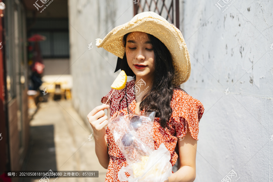 年轻漂亮的泰国亚裔旅行家，穿着红裙子，戴着红帽子，喜欢吃一块菠萝，并带有街头美食的概念。