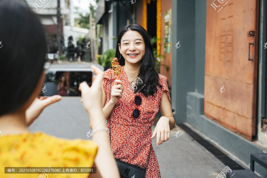 年轻漂亮的亚洲女人穿着红衣服在智能手机镜头前吃着街头食物。美食博客。街头小吃。