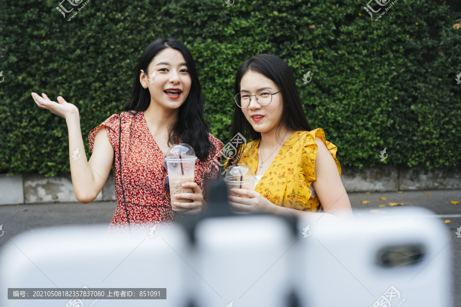 两位亚洲美女在自拍杆上的智能手机前摄像头前回顾饮料甜水。食物录影机-复制空间。