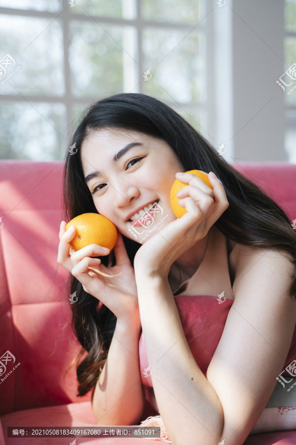 活泼的长发女孩趴在粉色沙发上，手里拿着橘子，笑容灿烂。