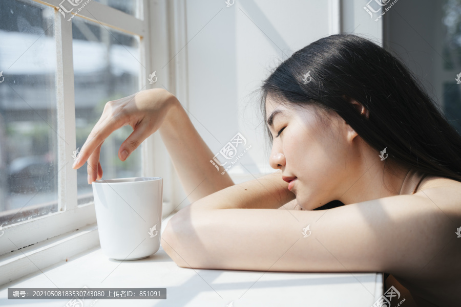 长发女孩坐在窗前，在晨光下端着一杯咖啡。