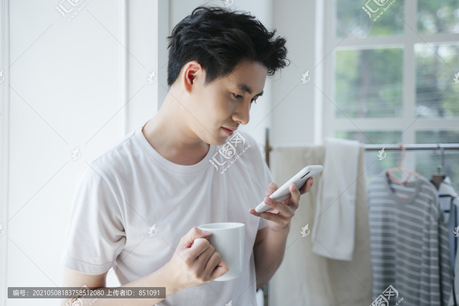 一个穿着白色t恤的男人拿着杯子，一边看手机，一边接听来电。
