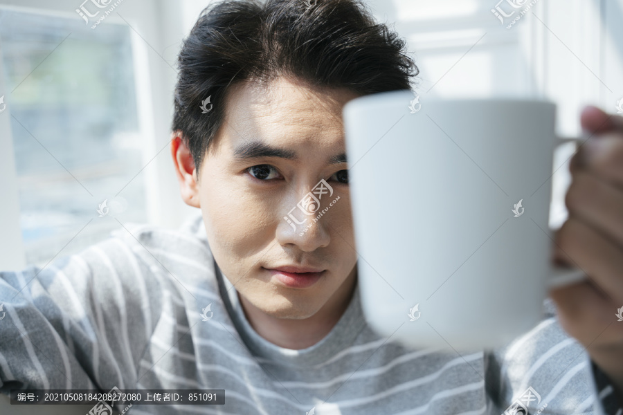 一个男人坐在床上，靠着窗户，端着一杯咖啡，阳光透过窗户。