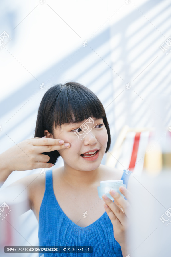 美丽的bob发型，穿着蓝色背心衬衫的亚洲女人，手里拿着奶油乳液瓶罐，在房间里往脸上涂奶油。
