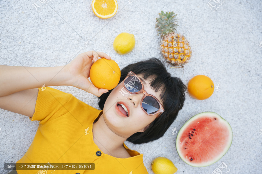 穿着黄色衬衫、戴着墨镜的亚洲年轻女子躺在地上，身上沾满了热带水果。