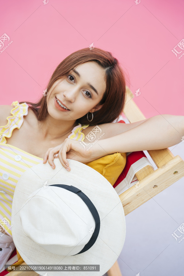 美丽的年轻女子拿着帽子，躺在沙滩上的椅子上。在粉红色背景上孤立。暑假。
