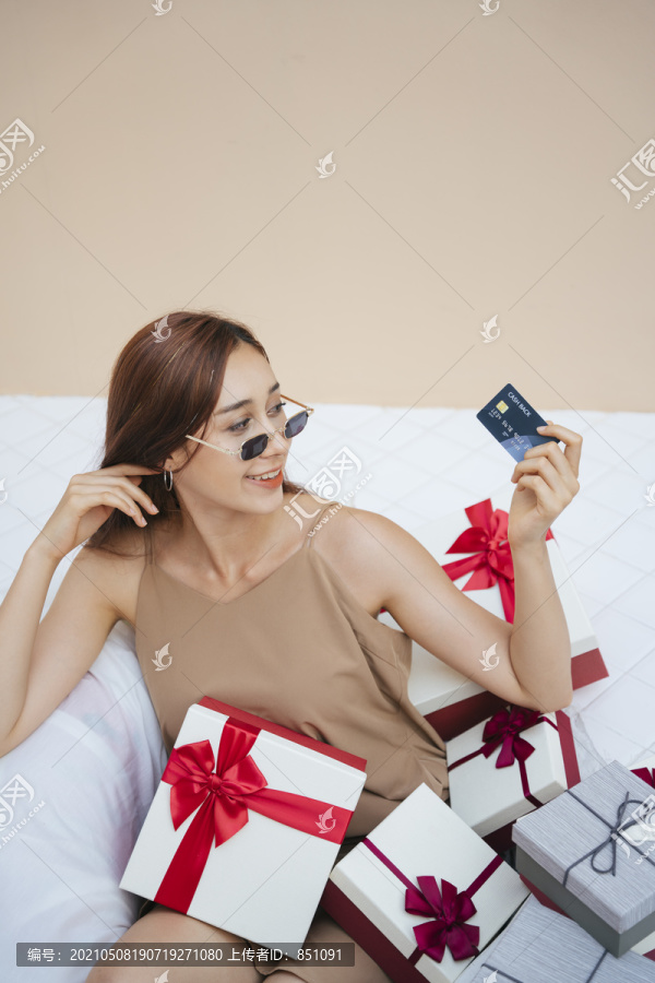 戴着墨镜的年轻漂亮女人坐在白色沙发上，手里拿着很多礼品盒和信用卡。假日网上购物。以米色奶油色为背景。