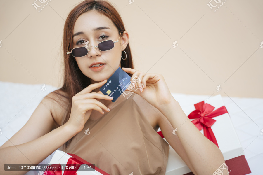 戴着墨镜拿着信用卡的漂亮女人，坐在白色沙发沙发上的礼品盒上。以米色奶油色为背景。
