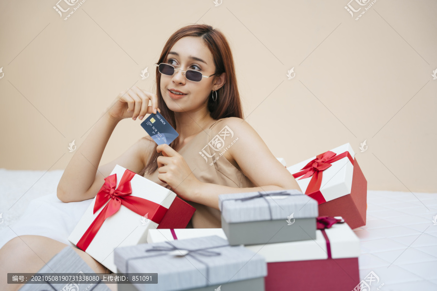 戴着墨镜拿着信用卡的漂亮女人，坐在白色沙发沙发上的礼品盒上。以米色奶油色为背景。