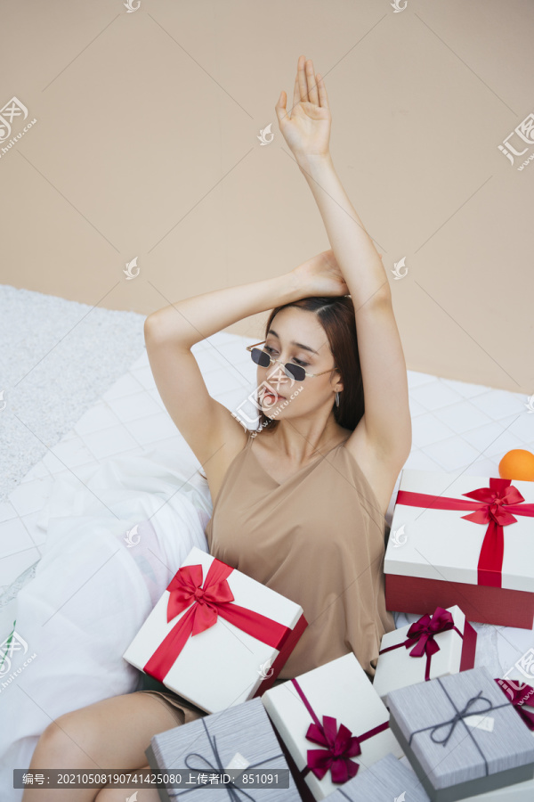 美丽的亚洲女人手举在空中戴着太阳镜坐在白色沙发沙发与礼物礼盒负荷。以米色奶油色为背景。