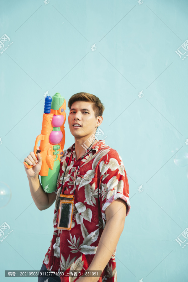 英俊的泰国当地男子身穿彩色花布手持水枪。隔离在柔和的蓝色背景上。泰国的泼水节。