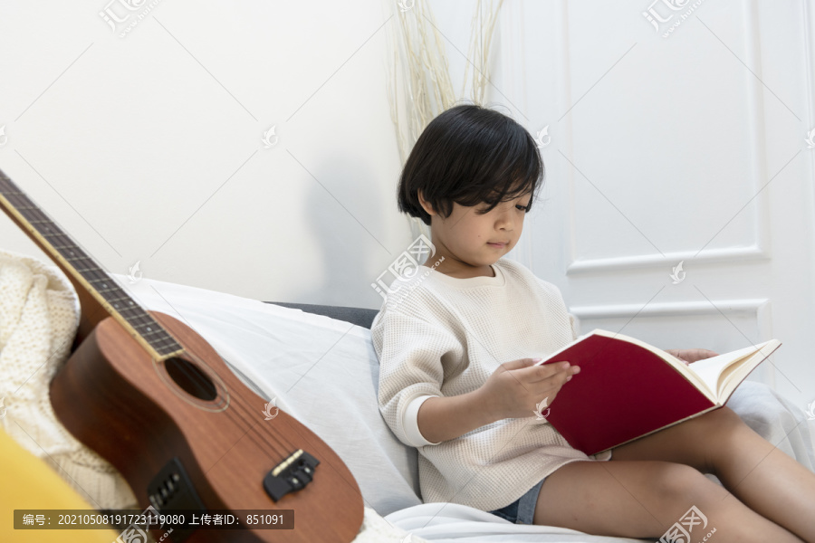 可爱的小男孩在读一本书的故事，坐在沙发上与四弦琴在客厅。阅读书中的吉他笔记。