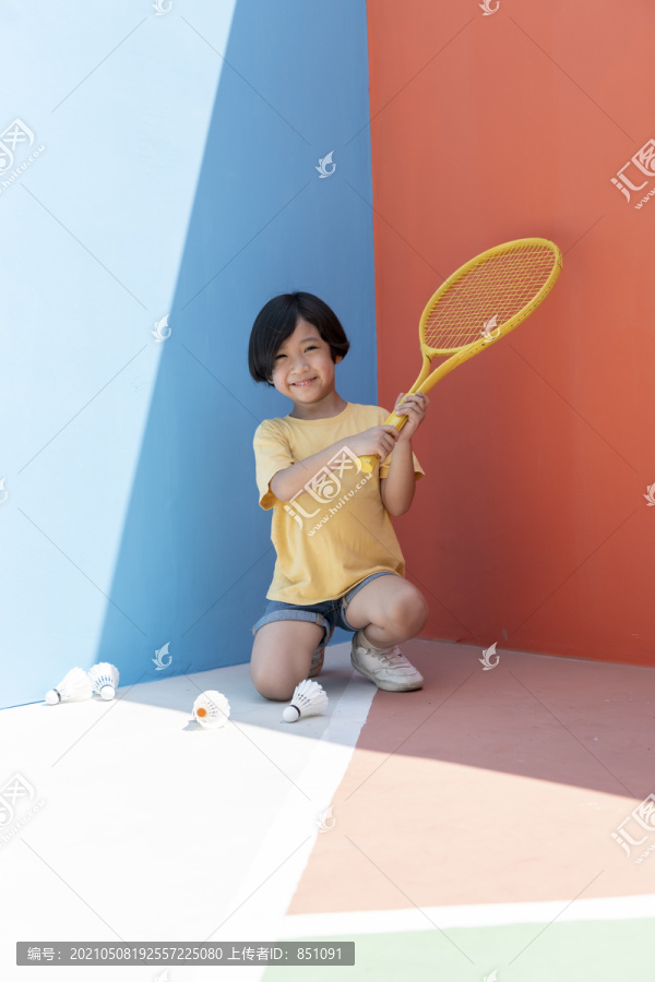 穿着黄色衬衫的可爱小男孩拿着网球拍，背景是五颜六色的蓝红色。地板上的羽毛球。坐在球场上。