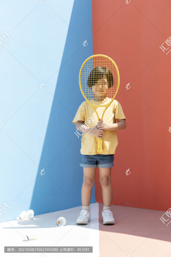 穿着黄色衬衫的可爱小男孩拿着网球拍，背景是五颜六色的蓝红色。地板上的羽毛球。网球拍后面的男孩。