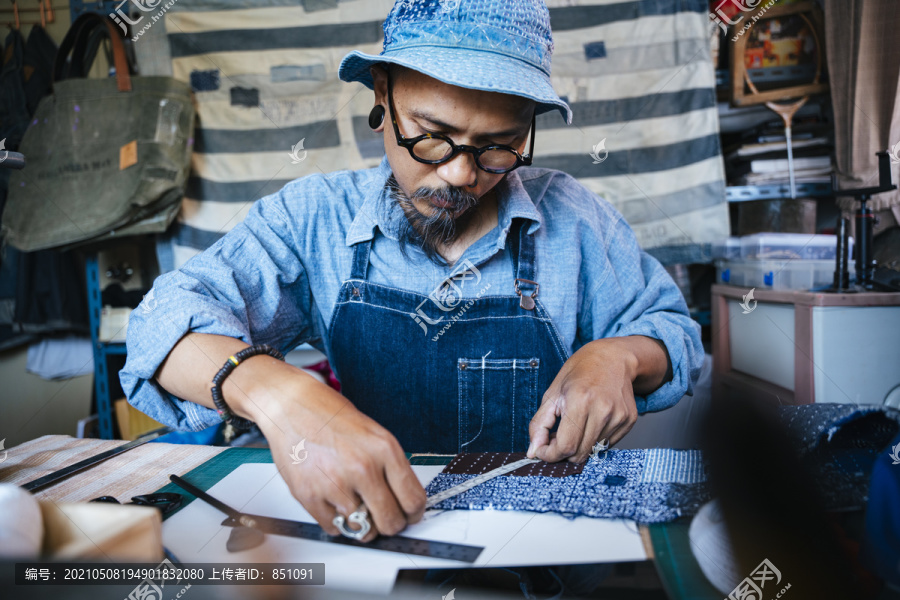 一个穿着蓝色衣服，戴着帽子和围裙的男人从纸上的图案剪下衣服。