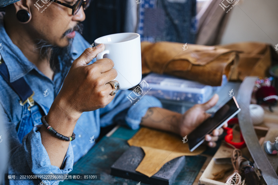一个穿蓝色衬衫的男人暂停他的皮革工作，喝咖啡来提神，在网上冲浪。