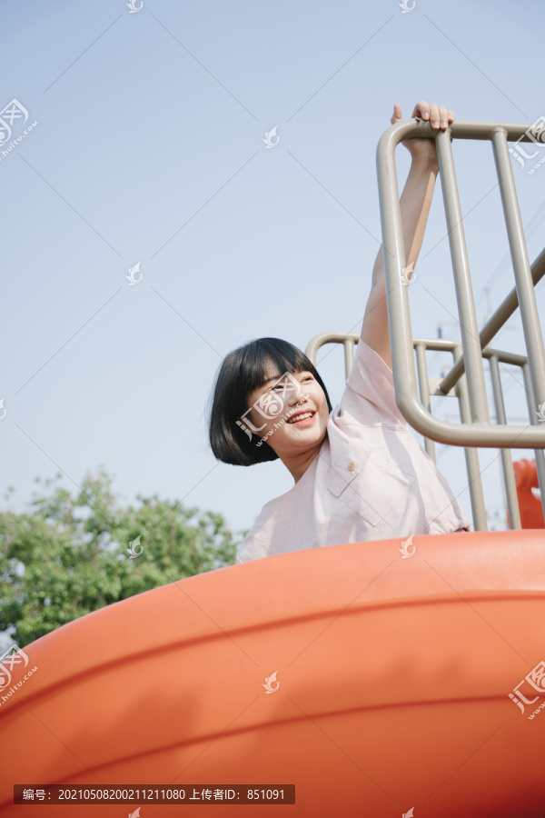 可爱的亚洲年轻女子鲍勃发型享受假期在游乐场公园坐在滑梯上。童年的快乐。