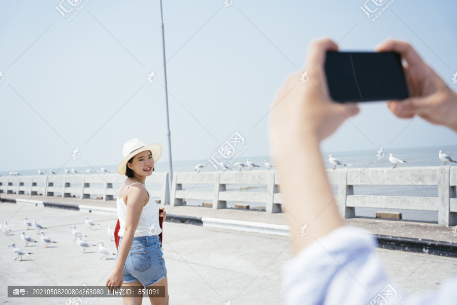 男人拿着智能手机的手拍着女人在海边旅行戴着帽子的女人。