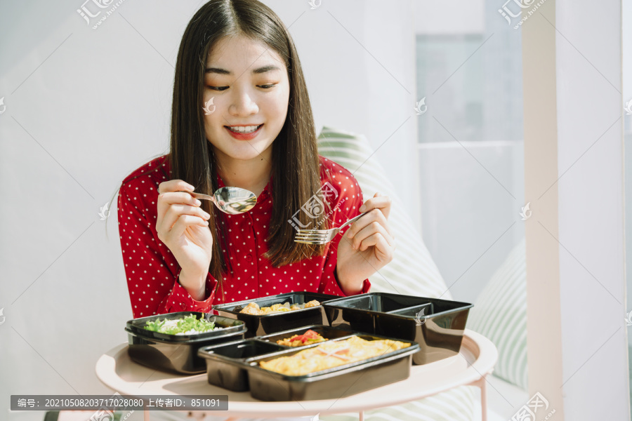 美丽的亚泰长黑发红衫女子拿着用叉子和勺子吃食物的饭盒套装。