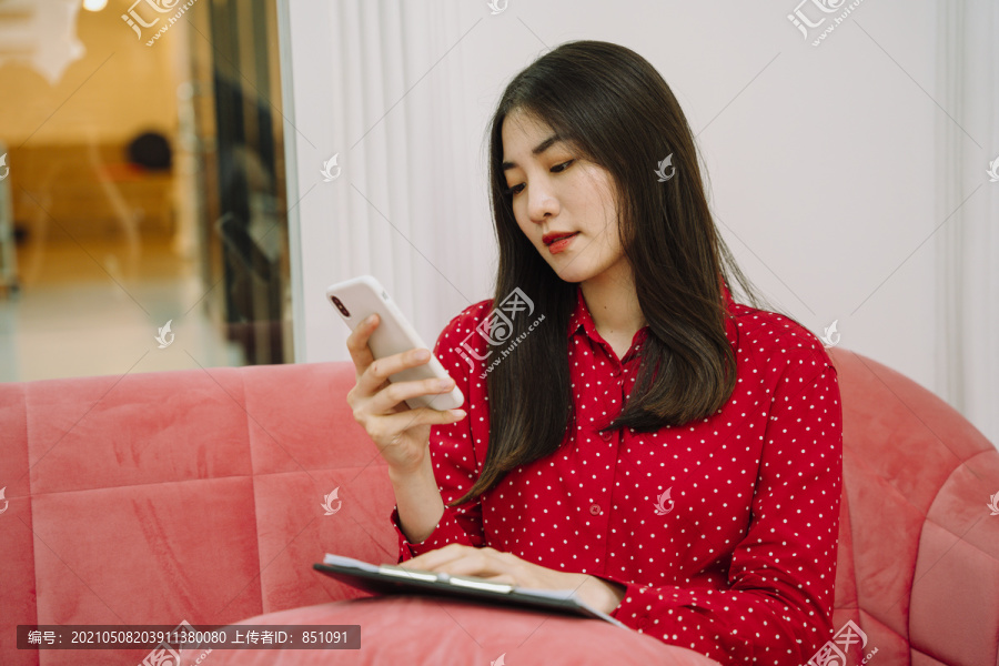 美丽的亚泰黑发红衫女子坐在粉色沙发沙发上，用智能手机与顾客联系，核对纸质工作清单。在家工作。在家工作