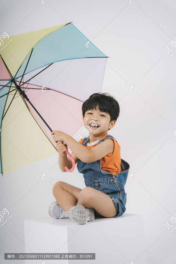 穿着牛仔裤工作服的可爱小男孩坐在工作室的白色街区上，手里拿着彩色粉彩伞。