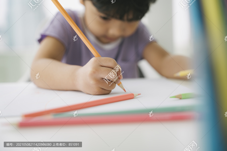 一个穿着紫色t恤的小男孩坐在靠窗的桌子旁，用铅笔和颜色在纸上画画和着色。