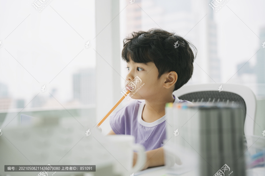 穿着紫色t恤的小男孩坐在靠窗的桌子旁，想着下一步要画什么。