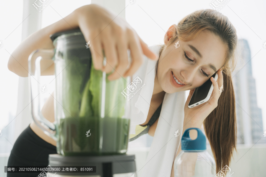 美丽健康的女人用搅拌机做健康蔬菜汁，在厨房里和朋友通电话。