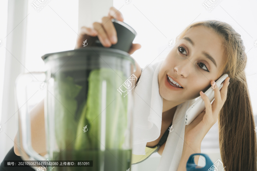 美丽健康的女人用搅拌机做健康蔬菜汁，在厨房里和朋友通电话。