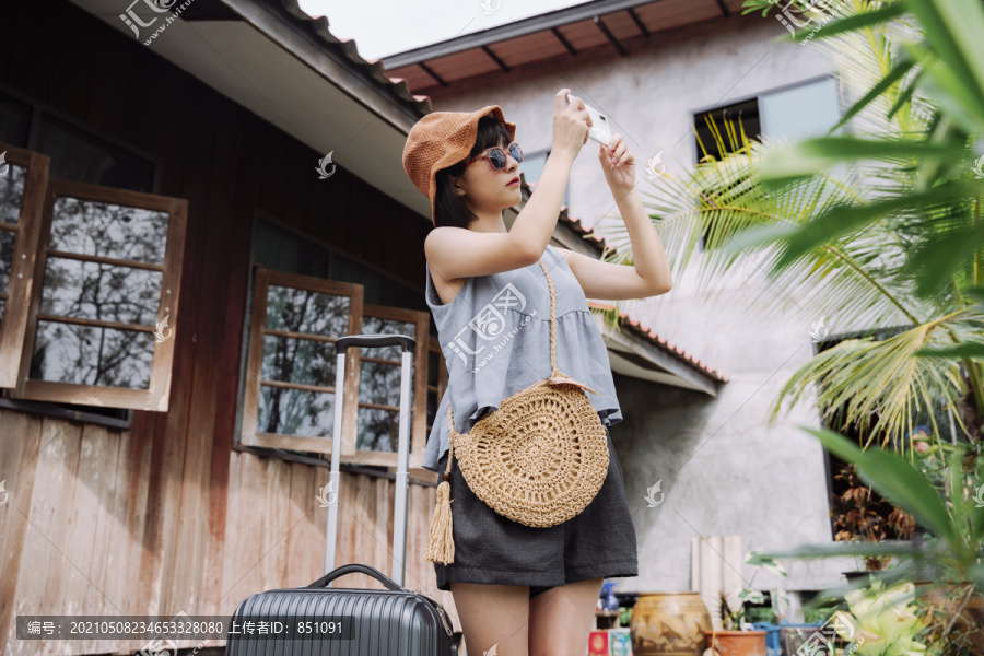 美丽的亚泰女旅行家，穿着夏季休闲服，戴着帽子，带着当地酒店的手推车行李。本地旅行。